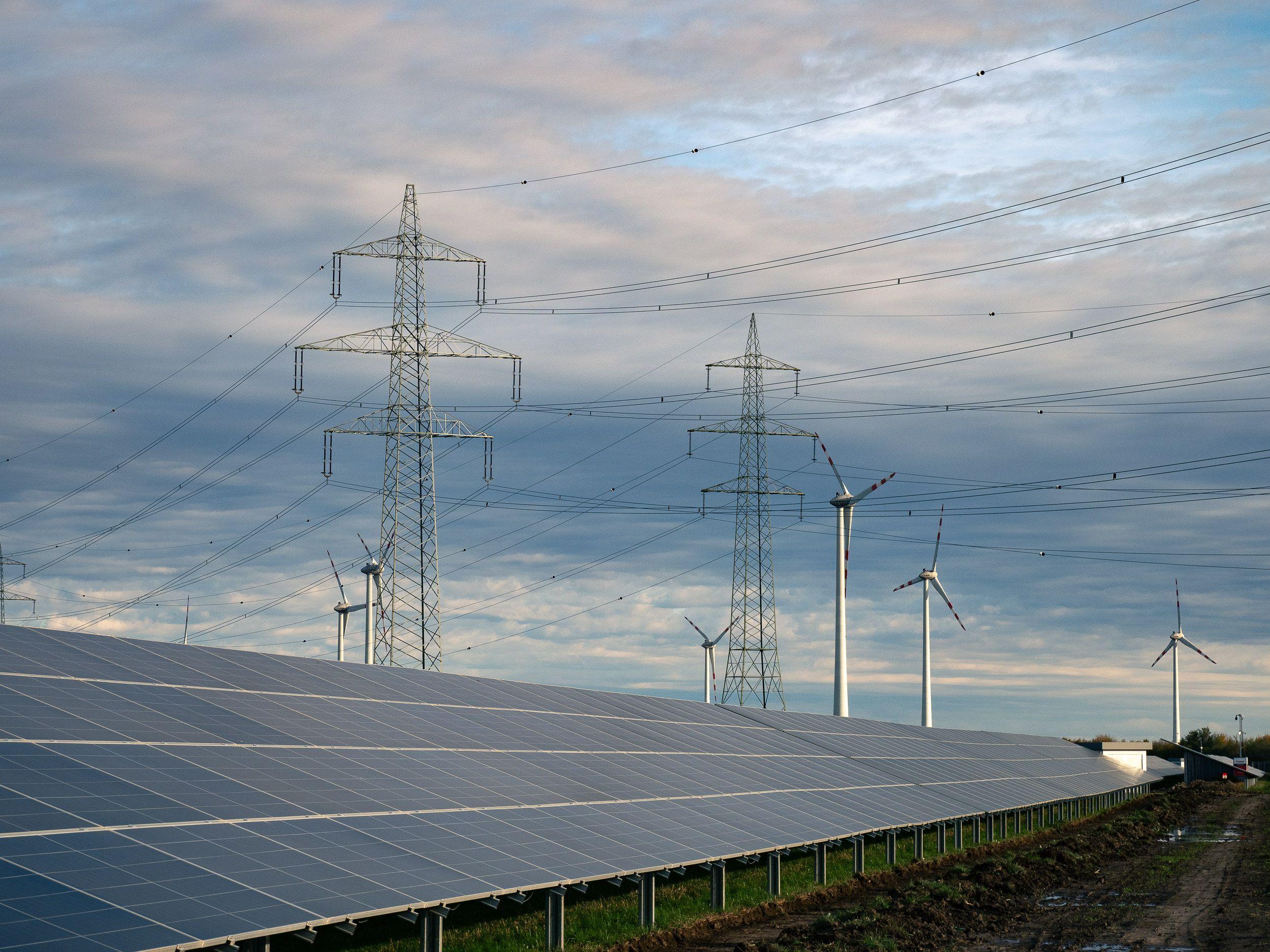 Photovoltaikmodule im unteren Bildabschnitt, Windräder und Strommasten erstrecken sich bis zum Horizont unter einem blauen Himmel mit Wolken.