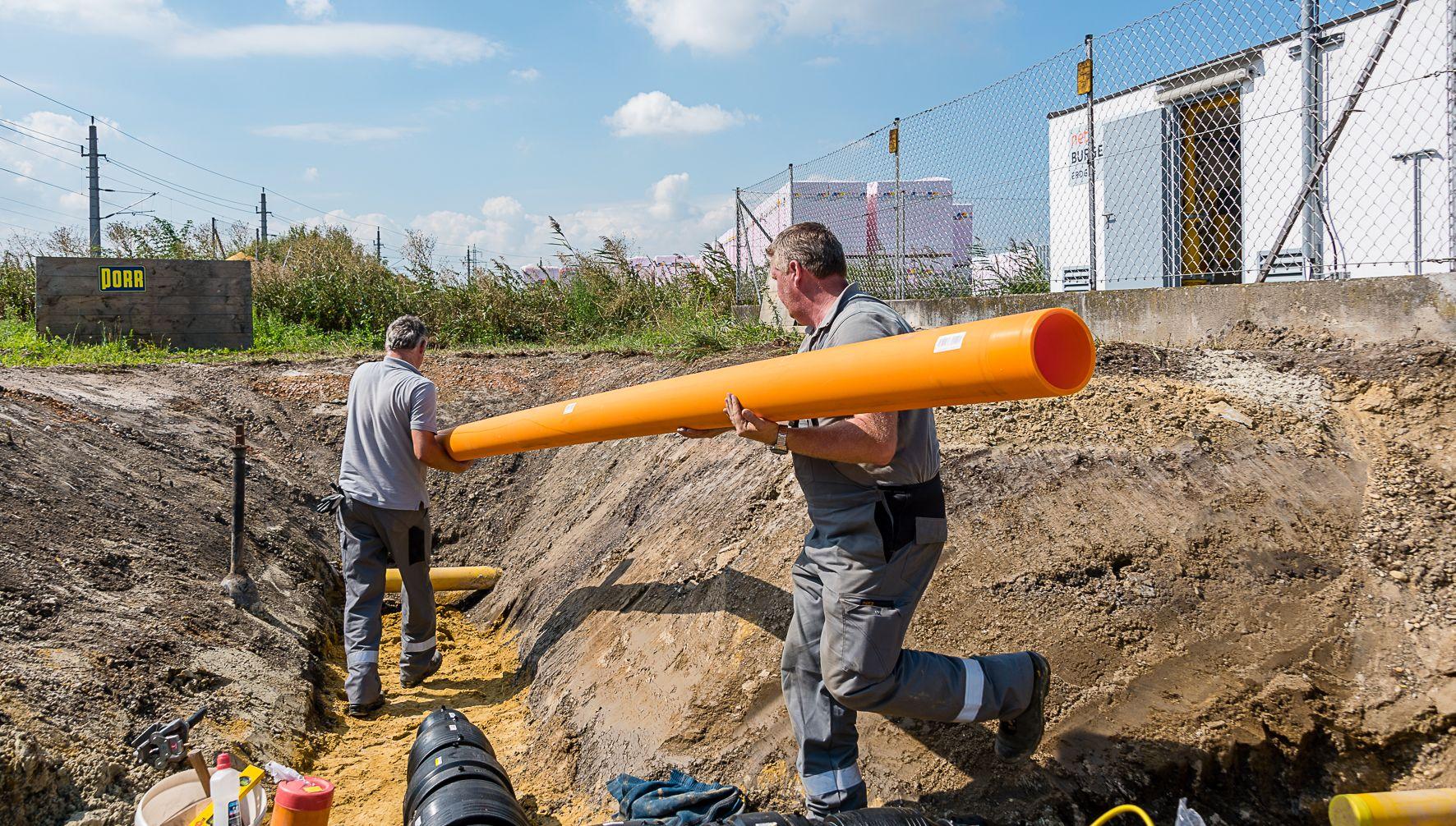 Zwei Techniker heben gemeinsam ein orange gefärbtes Gasrohr in eine Baugrube.